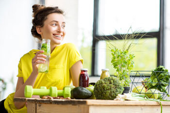 Eine junge Frau in einem gelben T-Shirt trinkt ein Getränk, während sie an einem Tisch sitzt, auf dem Gemüse und Hanteln liegen.