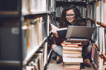 Studentin sitzt mit Bücherstapel und Notebook zwischen Bücherregalen auf dem Boden