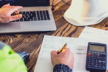 Student sitzt mit Laptop und Taschenrechner an Schreibtisch und notiert sich etwas