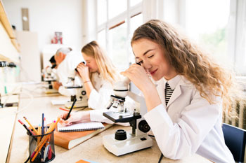 Zwei junge Frauen in Laborkitteln schauen in Mikroskope und machen sich Notizen an einem Schreibtisch