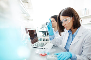 Eine junge Frau in Laborkleidung hantiert mit Pipette, Proben und Notebook, während ein junger Mann hinter ihr in ein Mikroskop schaut.