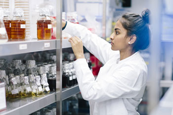 Eine junge Frau im Laborkittel greift nach Glas mit einer Flüssigkeit, das in einem Regal voller Flüssigkeitsbehälter steht.