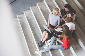 Vier Frauen sitzen mit Laptop, Smartphone, Tablet und Notizblock auf einer Treppe.