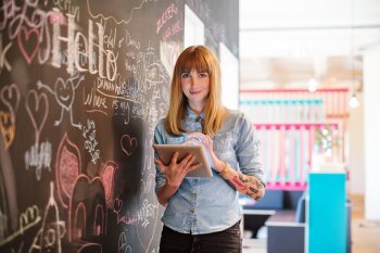 Design-Studentin steht mit Tablet vor bemalter Tafelwand und lächelt in Kamera