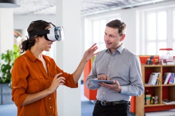 Junge Frau hat Virtual Reality Brille an, junger Mann steht daneben und macht sich Notizen