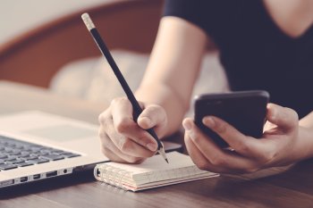Studentin sitzt mit Smartphone in der Hand an Tisch und macht sich Notizen