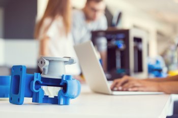 Im Vordergrund ein Plastikteil, im Hintergrund zwei Studierende an 3D-Drucker