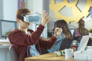 Junger Student sitzt mit Virtual Reality Brille am Tisch, im Hintergrund weitere Studentinnen