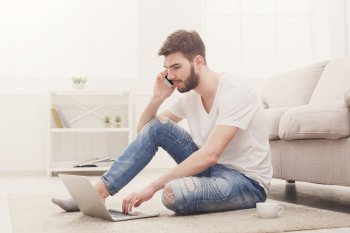 Junger Mann sitzt telefonierend mit Laptop auf dem Boden