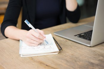 Junge Frau sitzt an Laptop und macht sich handschriftliche Notizen