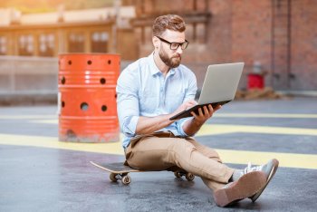 Junger Mann sitzt mit Laptop in der Hand auf einem Skateboard