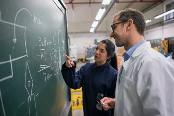 Zwei Studierende stehen in Kitteln an einer Tafel, auf der mathematische Formeln stehen