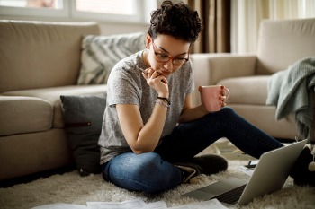 Eine junge Frau sitzt im Wohnzimmer auf dem Teppich, liest am Laptop und hält eine Kaffeetasse